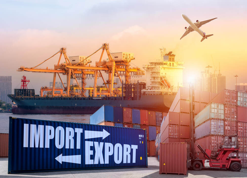  A container ship is being loaded with cargo at a busy port while an airplane flies overhead. In the foreground, a large container with the words 'import' and 'export' on it is being transported by a forklift, with more containers stacked behind it. In the background, a large ship is docked at a pier and a gantry crane is loading containers onto it.